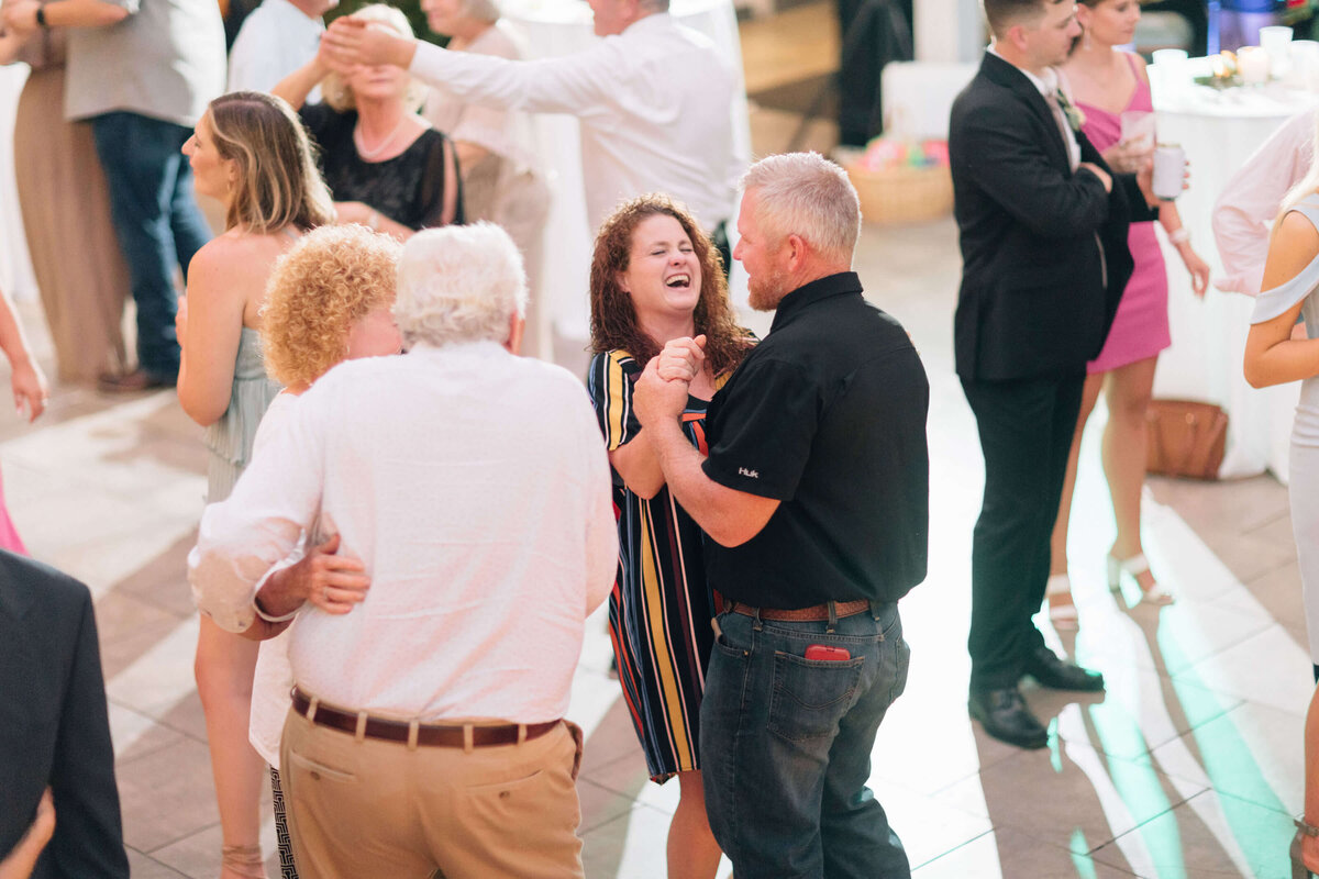 A group of people dance at a lively outdoor event. In the center, a woman in a striped dress smiles and laughs while dancing with a man in a black shirt. Others chat or dance in the background, creating a joyful atmosphere.