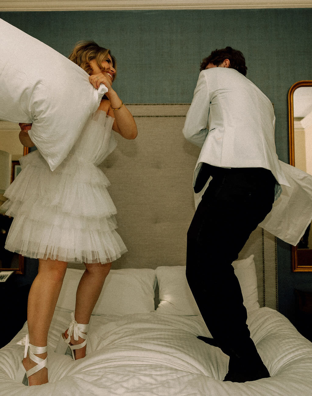 A newlywed couple standing on a bed having a pillow fight.