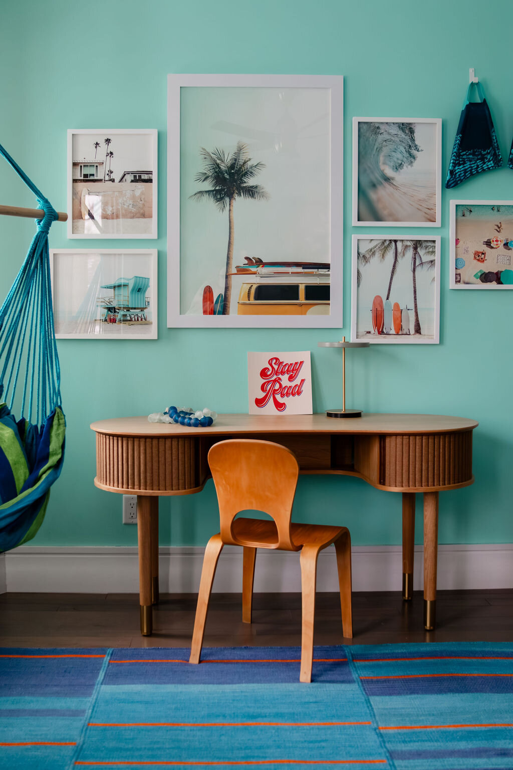 A bright, light turquoise room features a light-colored Umage Audacious desk with tambour doors and slender legs, positioned against a wall adorned with a gallery of framed beach-themed prints. These include images of palm trees, surfboards, and ocean waves. A small lamp and a postcard that reads "Stay Rad" sit on the desk. A simple wooden chair is placed in front of the desk. To the left, a blue and green hammock chair hangs near a window partially visible behind patterned curtains. A colorful striped rug lies on the dark wooden floor. Teal swimsuits hang on the wall to the right of the framed prints.