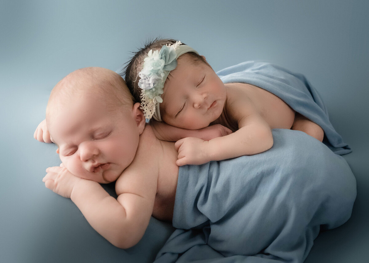 Newborn twins sleep on top of each other under blue blankets in a studio
