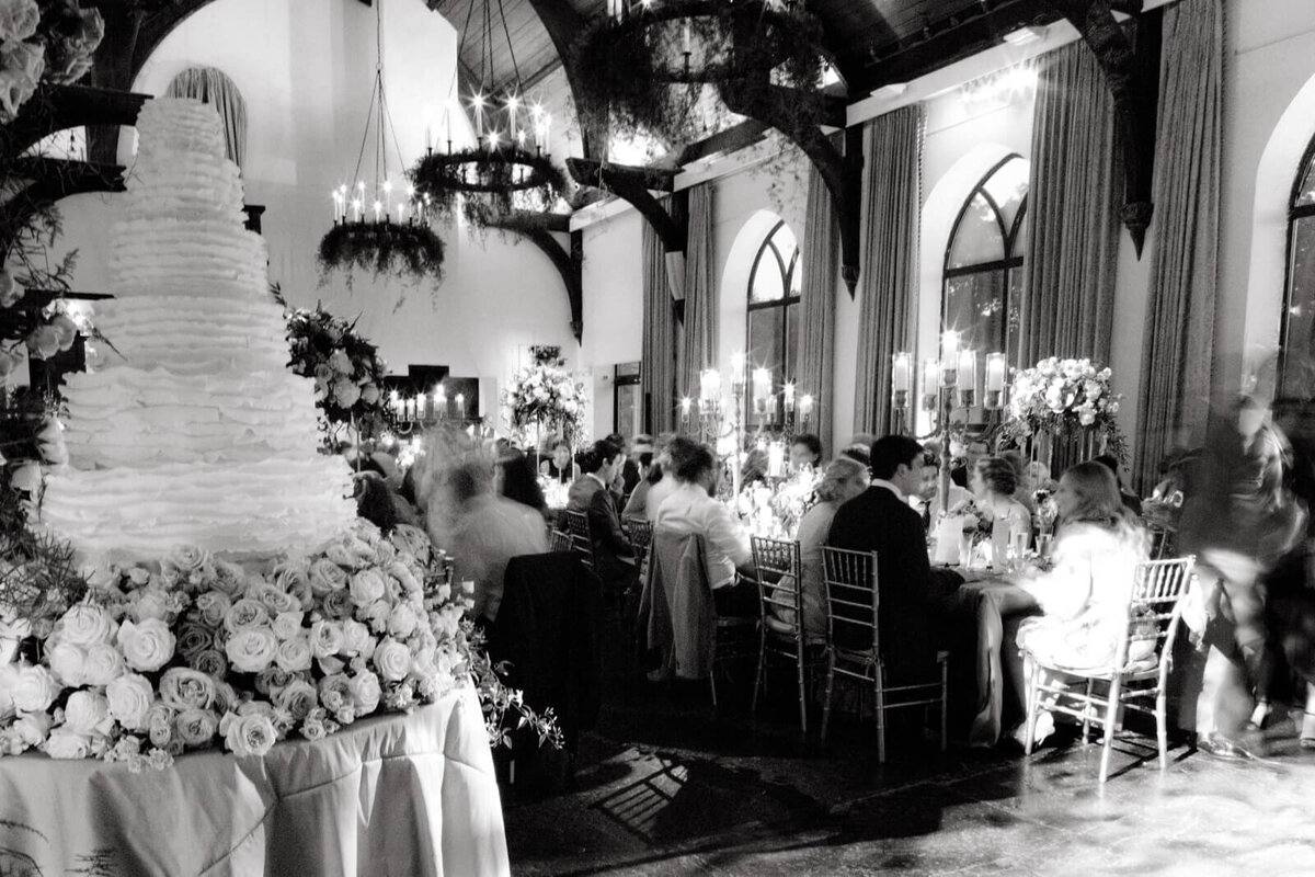Black and white photo of a large cake and a group of people chatting with each other on a long dining table.