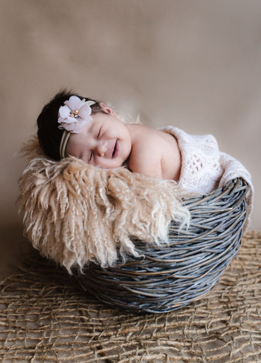 Smiling newborn baby with a light pink flower headband, resting on a fluffy beige blanket inside a rustic woven basket.