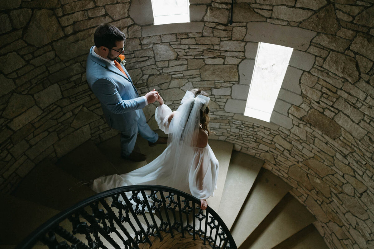 A bride and groom holds hands and walk down a round stairwell at the Chateu De Lis wedding venue.