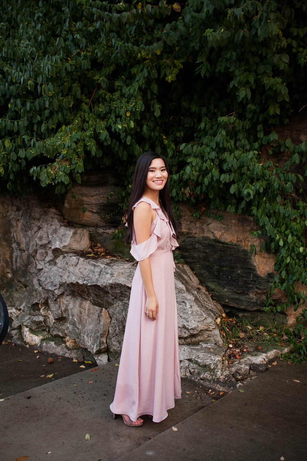 A lovely Asian American girl in a light pink dress with ruffles standing in front of a rock wall and overhanging trees. Captured by Springfield, MO senior photographer Dynae Levingston.