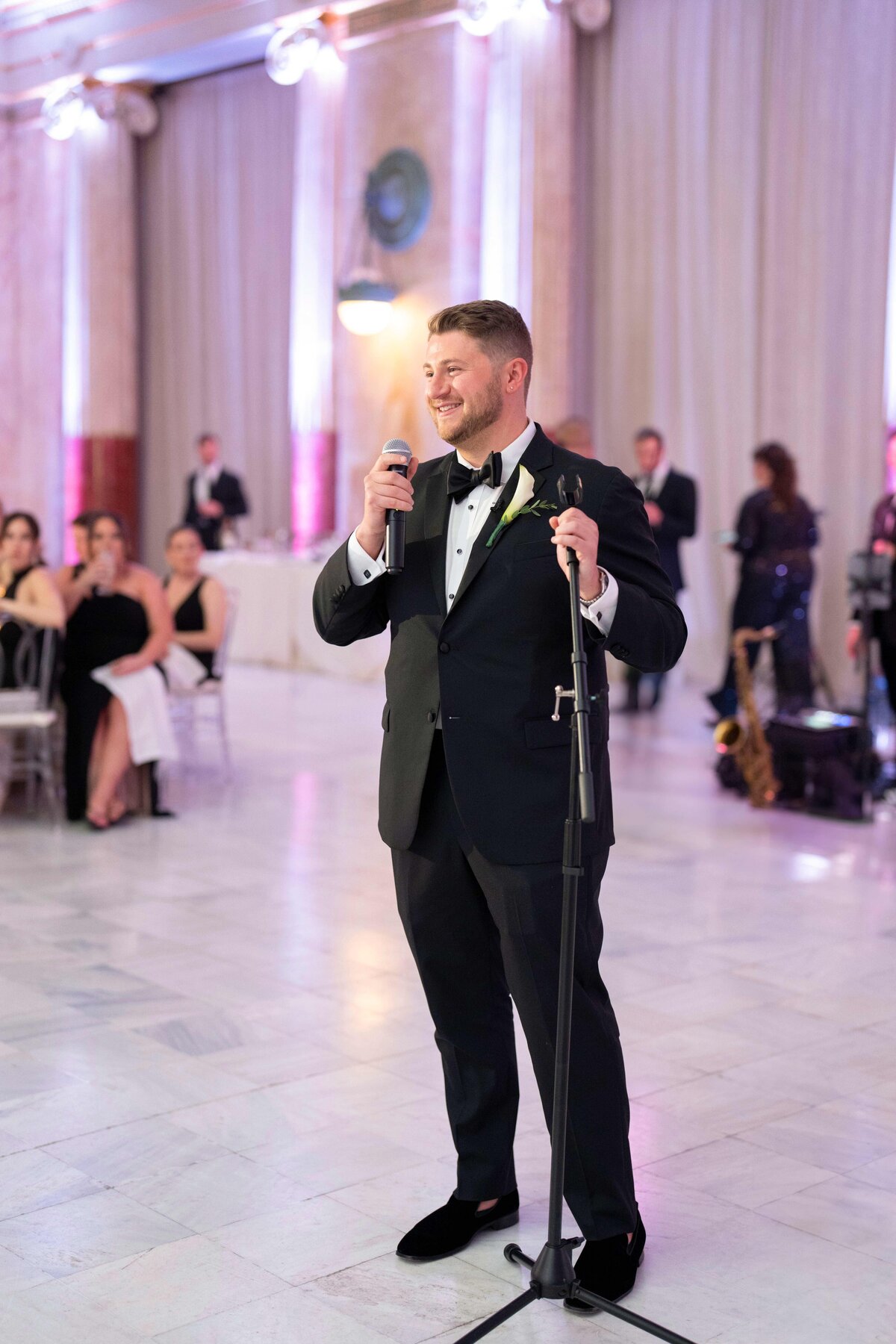 A man in a tuxedo stands smiling, holding a microphone, in a large, elegantly decorated room. People sit at tables in the background, listening. The room features marble columns and soft lighting.