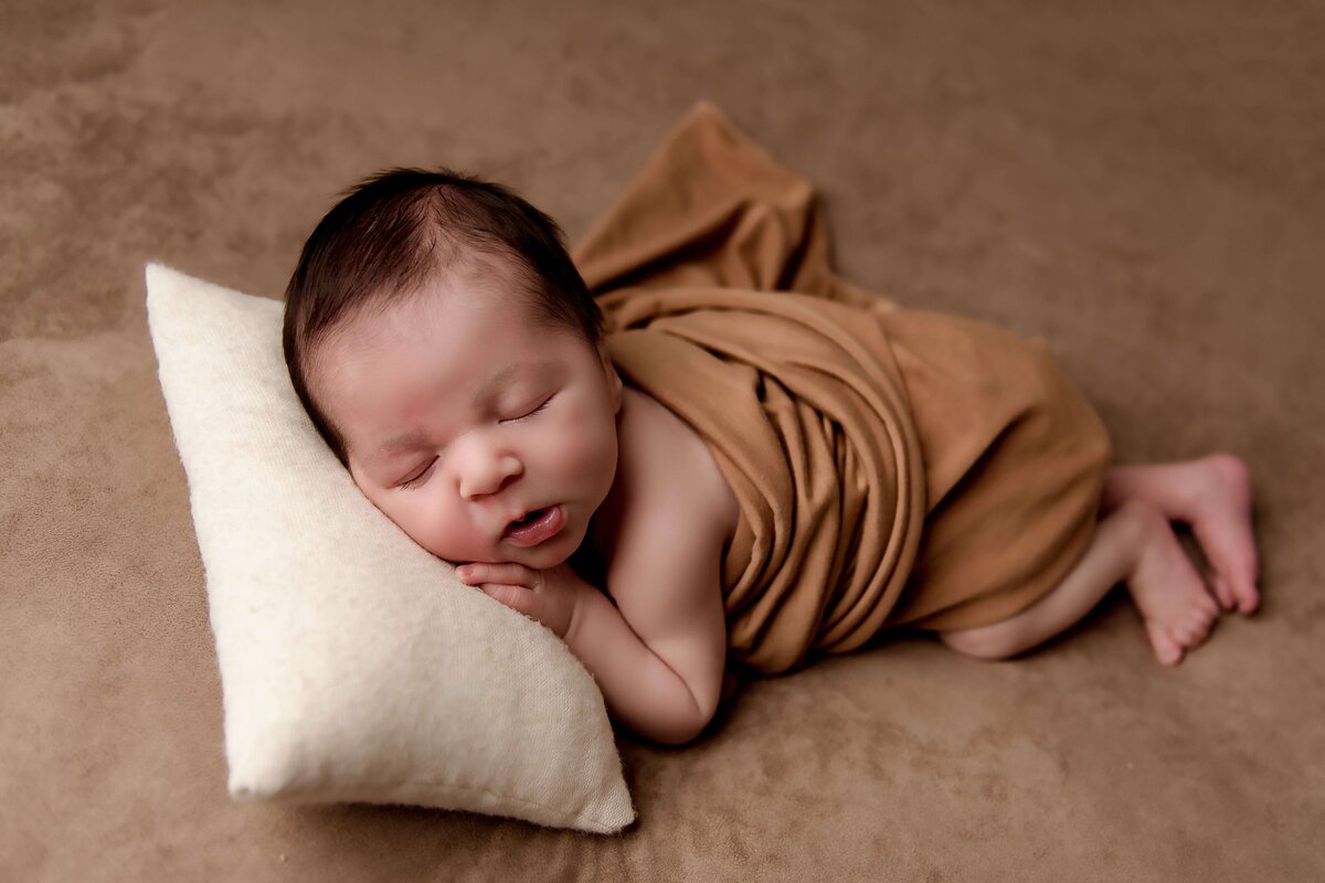 A newborn baby boy, resting his head on a cozy pillow for a perfect newborn portrait by Mobile, AL newborn photographer. The pillow is cream in color, with the blanket a cozy brown color, which matches the swaddle used to keep the baby warm.