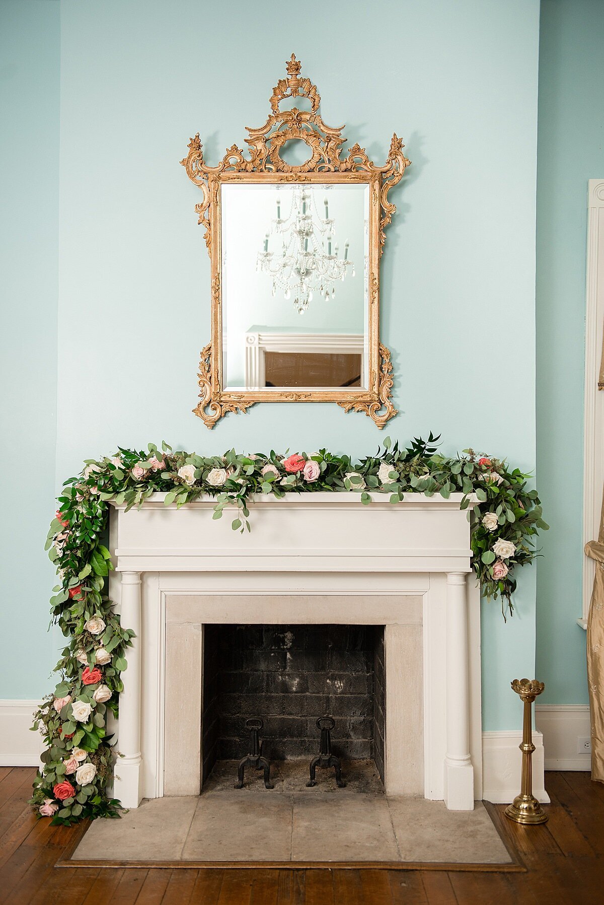 The large gilded gold mirror hangs above an ivory wood and marble mantle in the blue front room at Ravenswood Mansion. The crystal chandelier is reflected in the gold mirror, The mantle is decorated with a lush garland of greenery, accented by peach, blush, white and ivory rises and flowers.