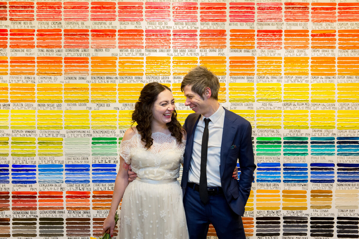 A bride and groom with their arms around each other in front of a colorful wall.