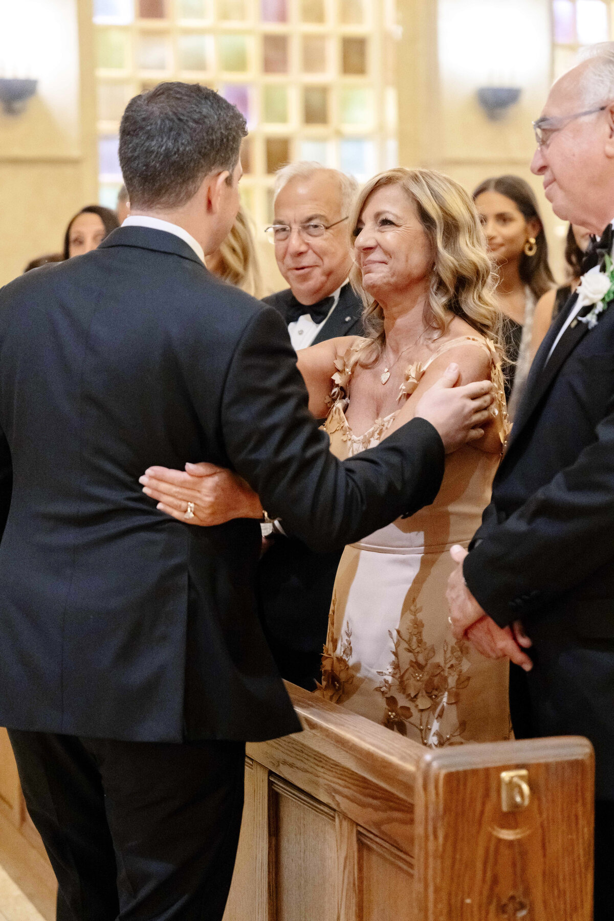 A groom, dressed in a black suit, warmly embraces a woman in a beige dress inside a church. Nearby, two older men in tuxedos watch with smiles. The background features soft lighting and people seated in pews.