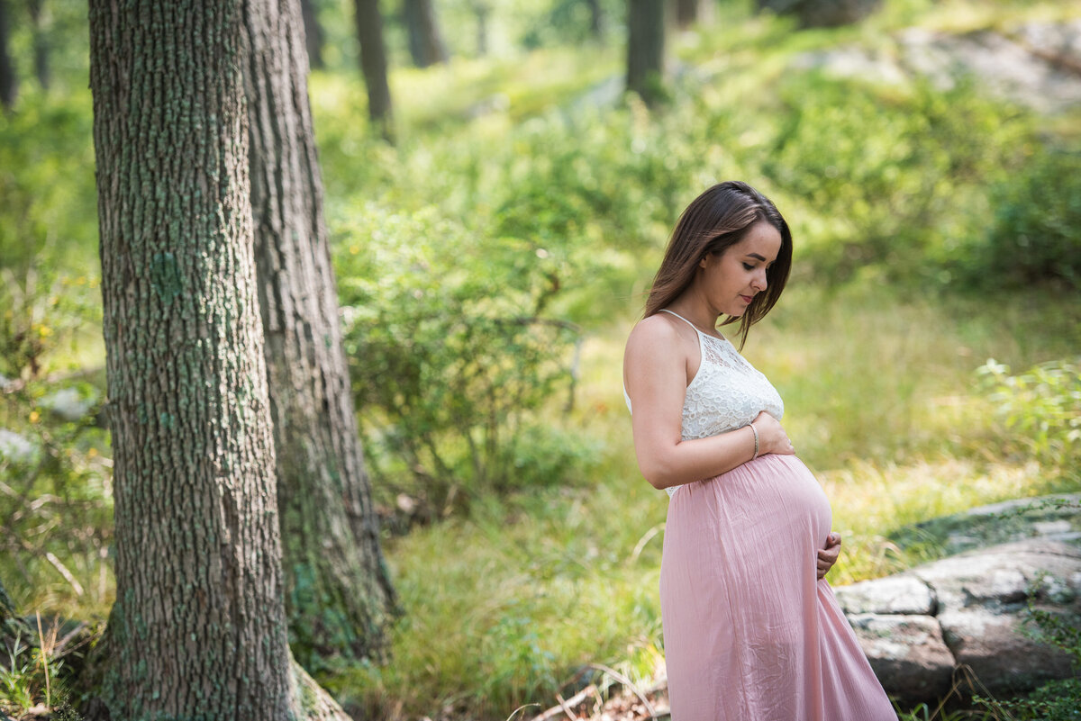 A person standing in a wooded area holding their pregnant stomach