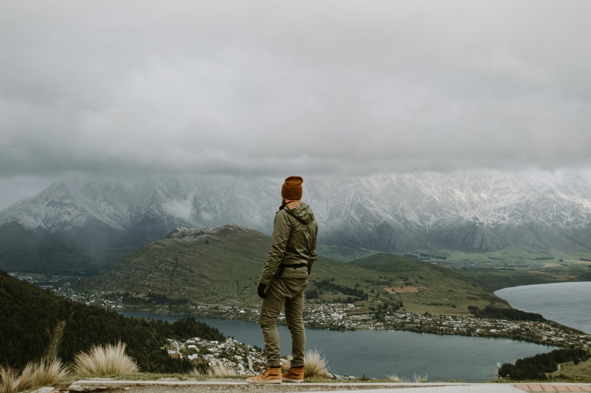 A man standing on a hill looking out over Queenstown towards a snowy Remarkables Mountain range.