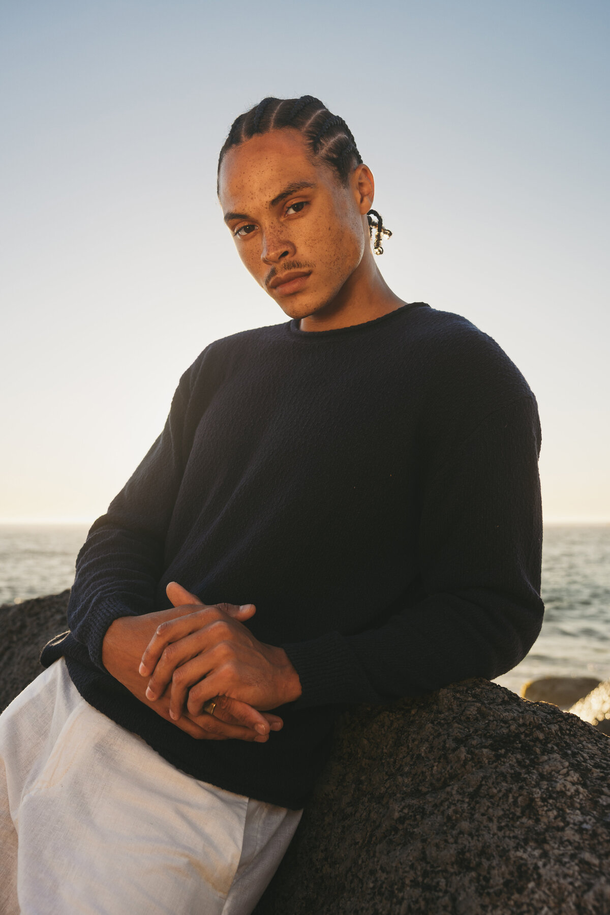 Portrait of male model Timothe leaning against a rock by the ocean at sunset in Cape Town, wearing a black sweater and white trousers