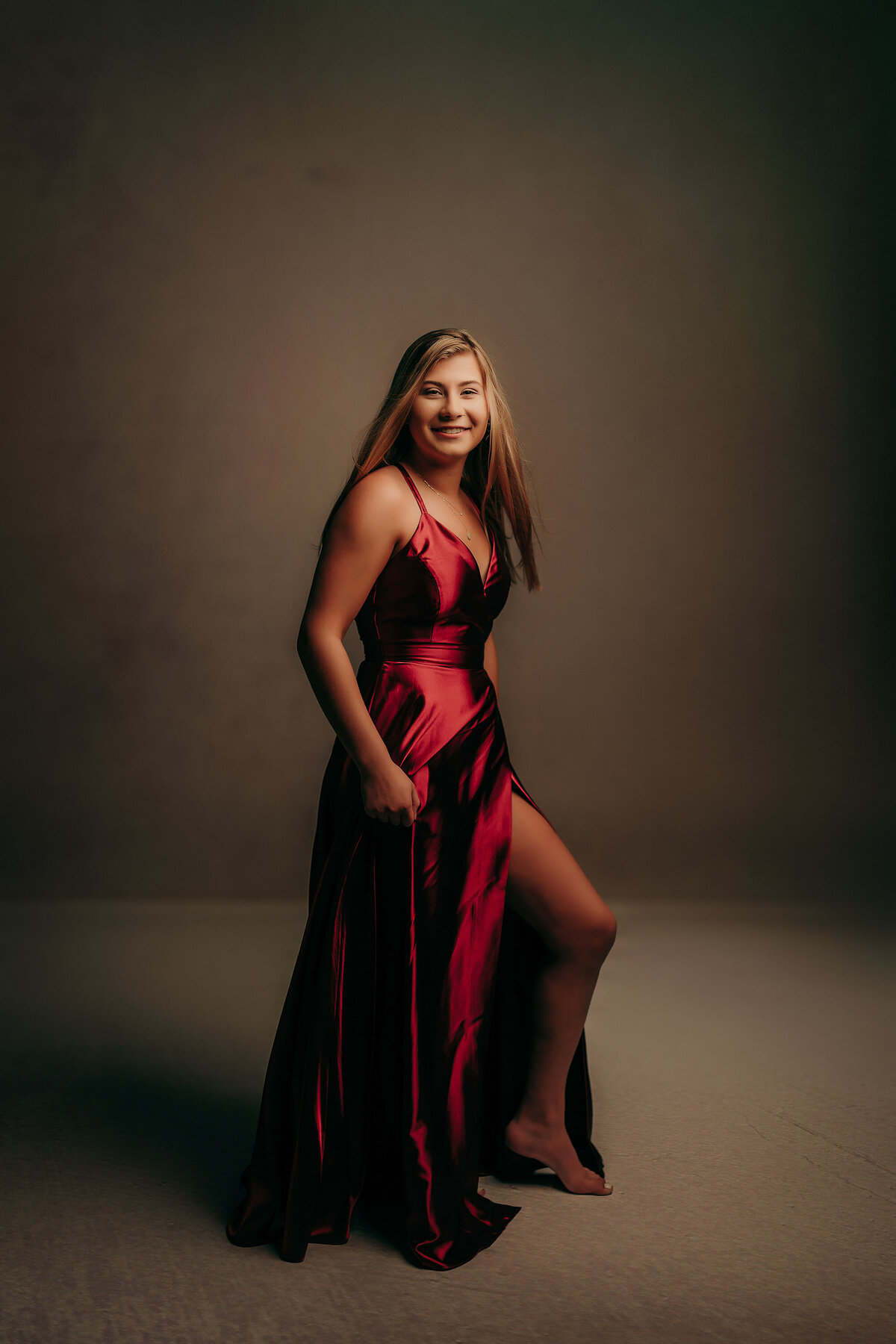 A Catholic Memorial High School senior stands in our Waukesha photo studio wearing a long, red, satin gown while posing for her senior portrait.