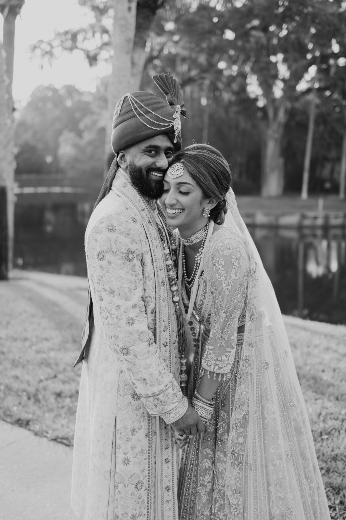 South Asian bride and groom dressed in traditional embroidered wedding attire share a joyful embrace, smiling warmly at the camera.