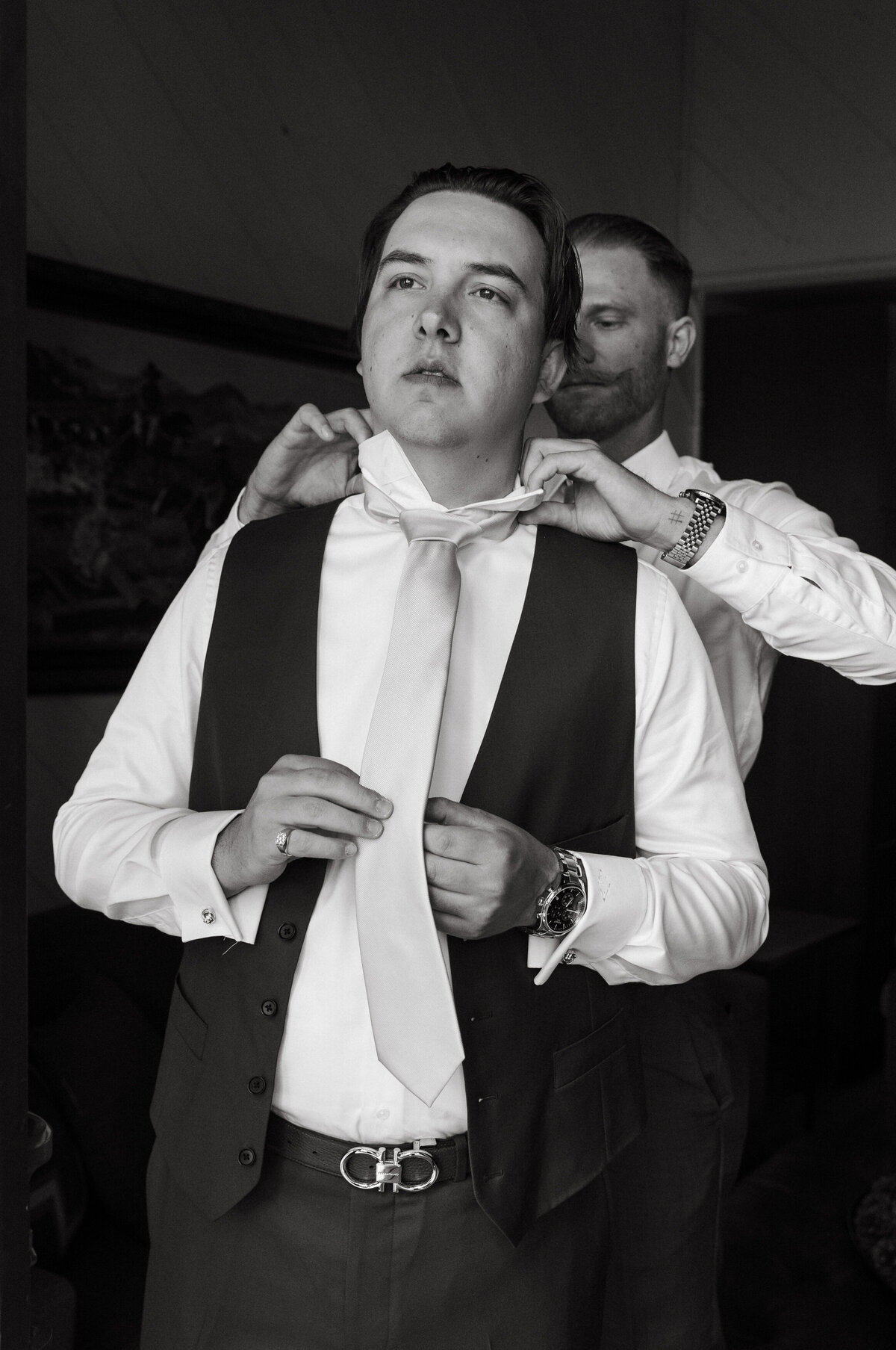 Black and white image of a groomsman helping the groom with his tie for his wedding.