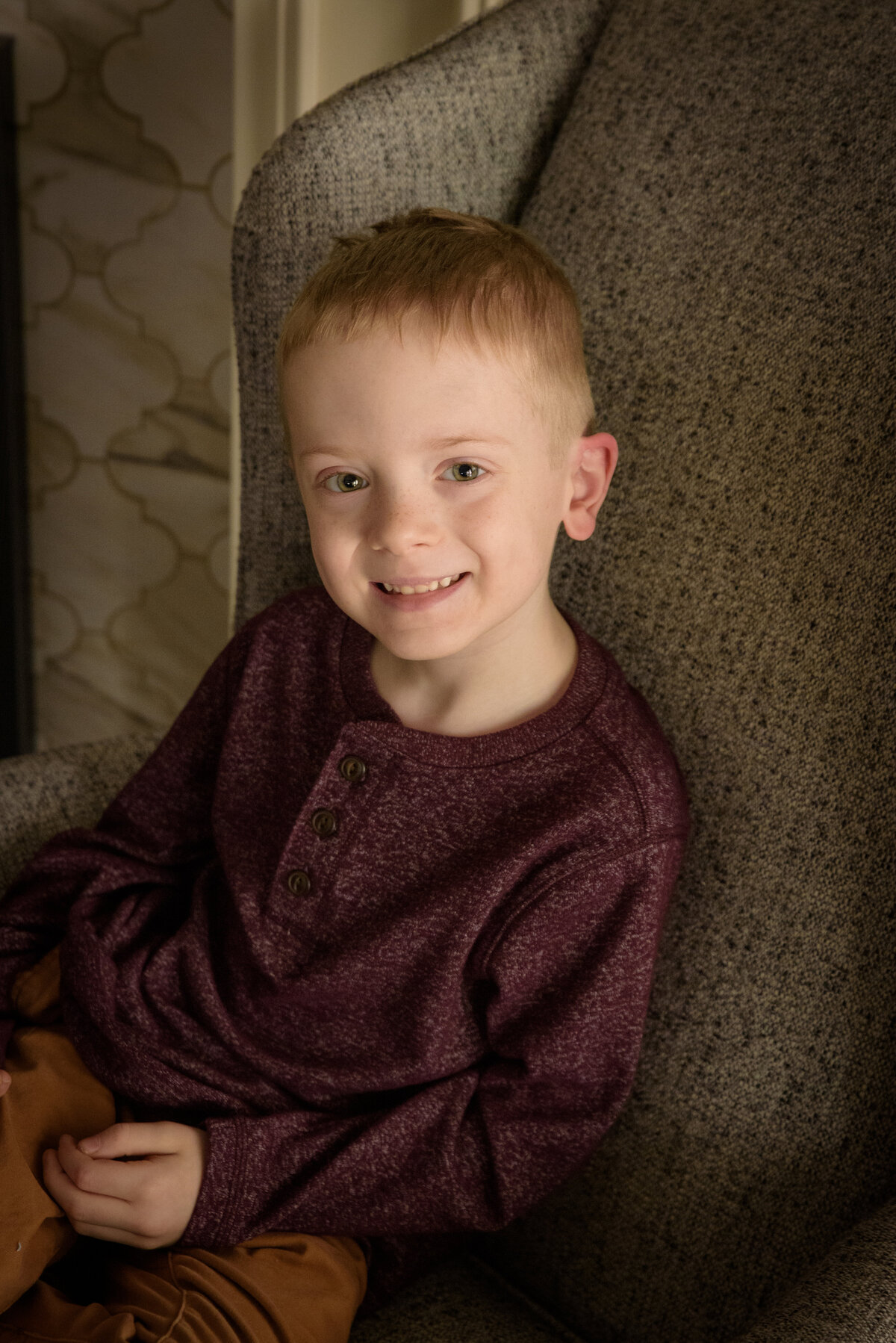 Natural light portrait of young boy wearing maroon shirt sitting in chair in his home in Green Bay, Wisconsin.