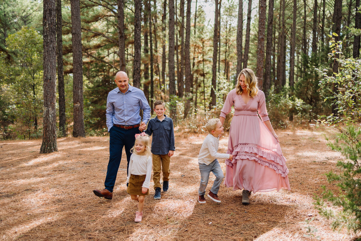 Family photo captured in a beautiful Albuquerque forest with tall trees and lush green leaves. The woman, wearing a light pink dress, is holding the hand of a child who is playing. The man, dressed in a dark blue shirt, is guiding a child in a light blue shirt by hand. In front of them, a blonde girl in a brown skirt and white blouse walks.