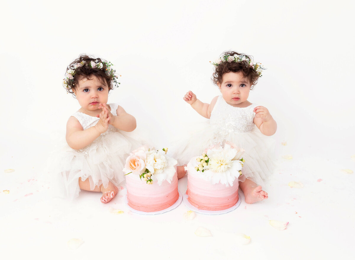 Timeless twin cake smash session with a pink and white setup, featuring soft pastel decorations and custom cakes. Capturing the joy and bond of twins celebrating their first birthday in a serene and elegant NYC photoshoot.