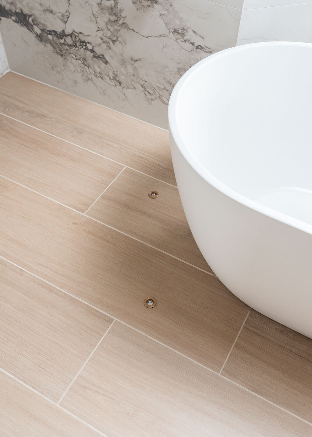 Close-up view of the corner of a modern white bathtub in a modern, grey marbled bathroom with light wooden floors.