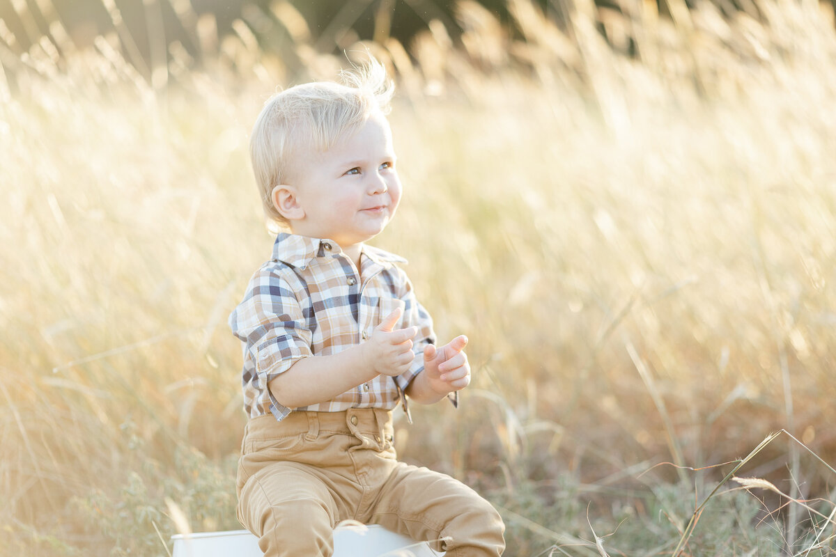 A cute little boy sitting in the grass as he looks off and smiles at his mother as they take family photos.