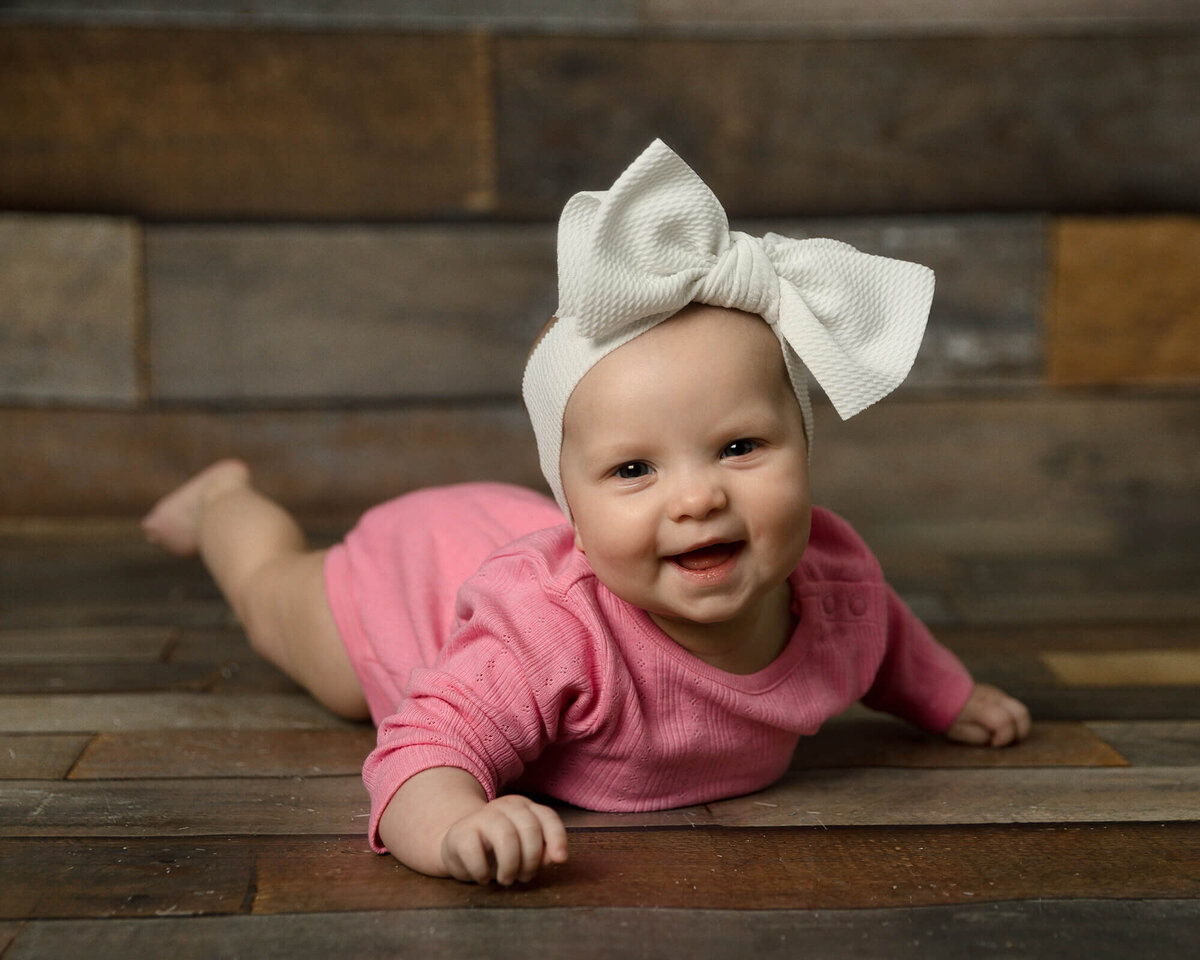 Baby girl in a dark pink outfit and large bow headband laying on her stomach. Wood background and floor.