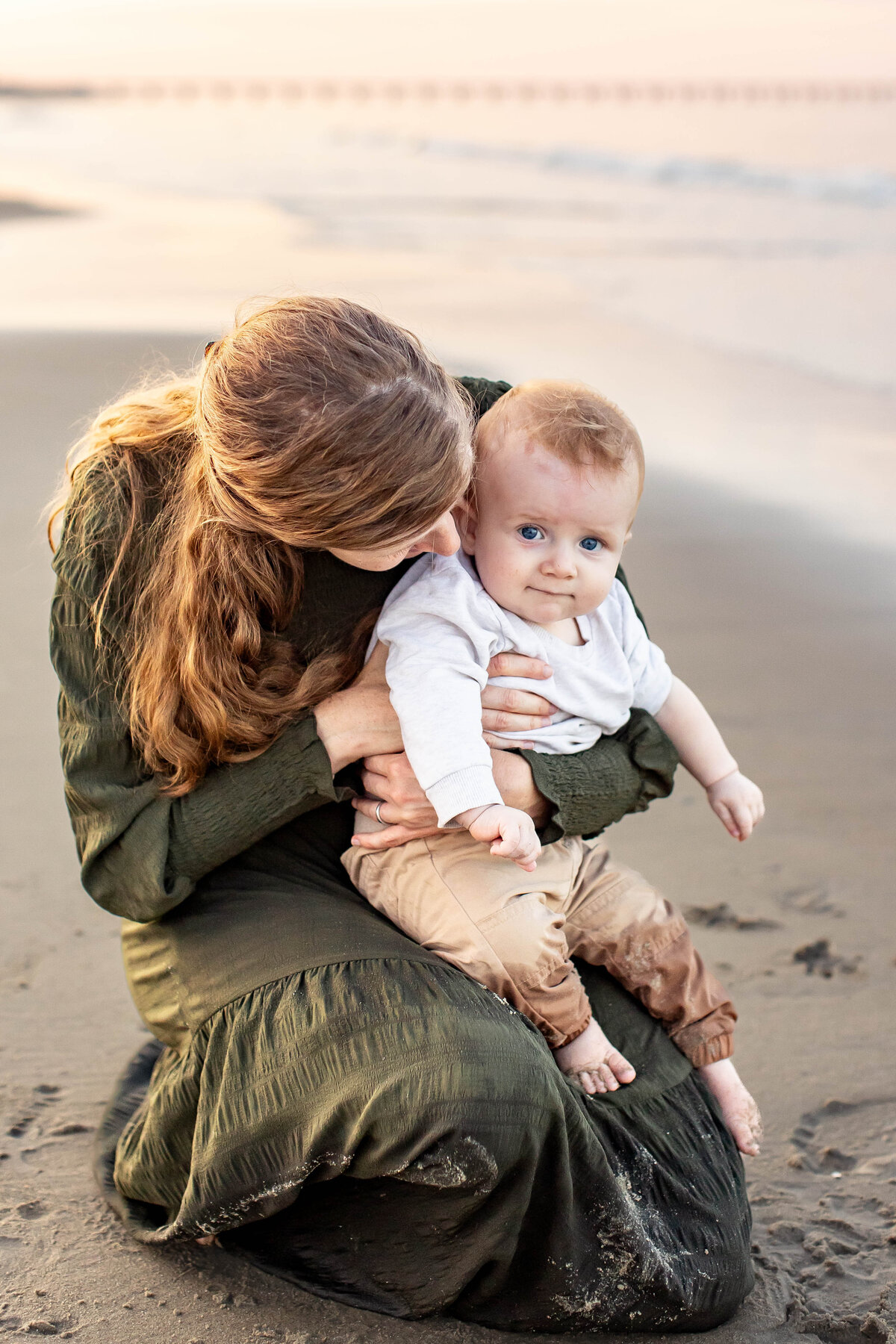 A mother lovingly holds her baby on the beach in Hampton Roads, Virginia, their hair tousled by the wind as they share a quiet, tender moment during a candid, playful motherhood session with Brooke Tucker Photography