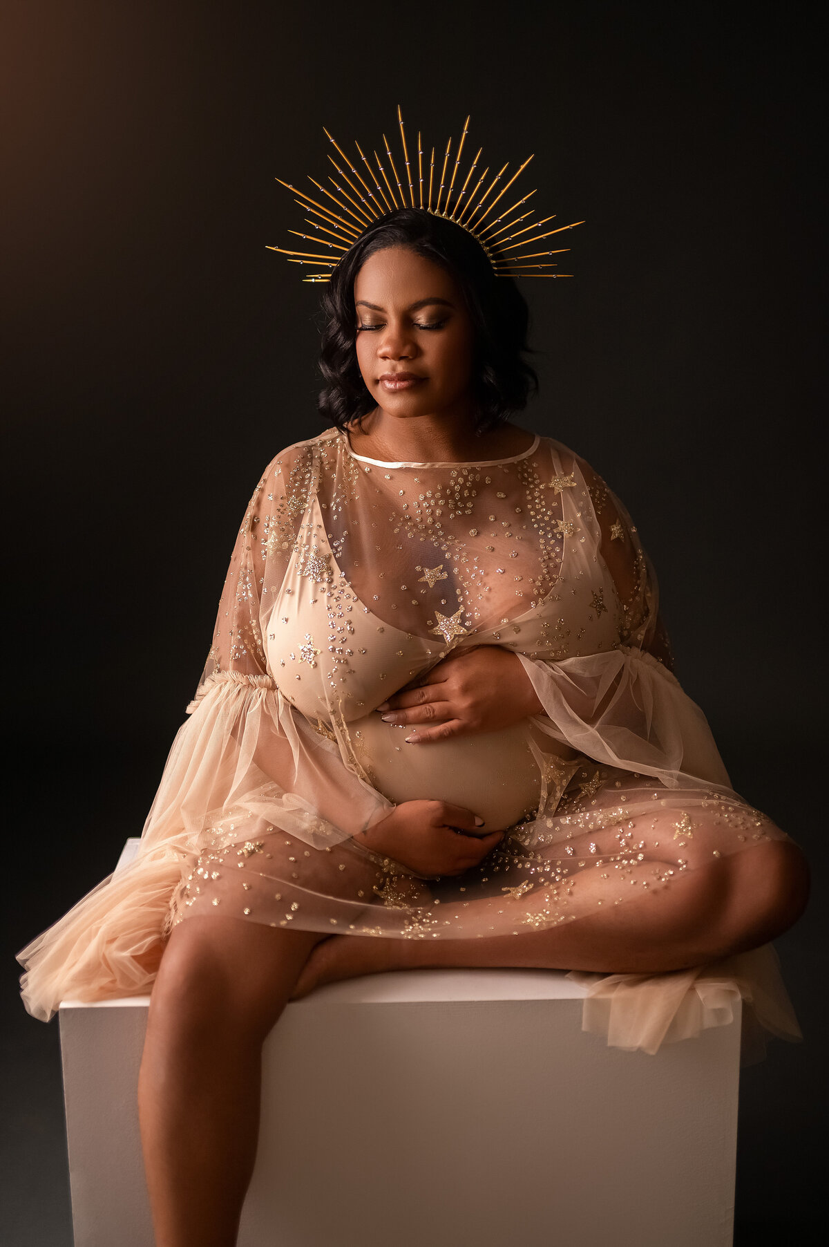 Pregnant Black woman in gorgeous shimmery gold bodysuit and crown, sitting upon a stool.