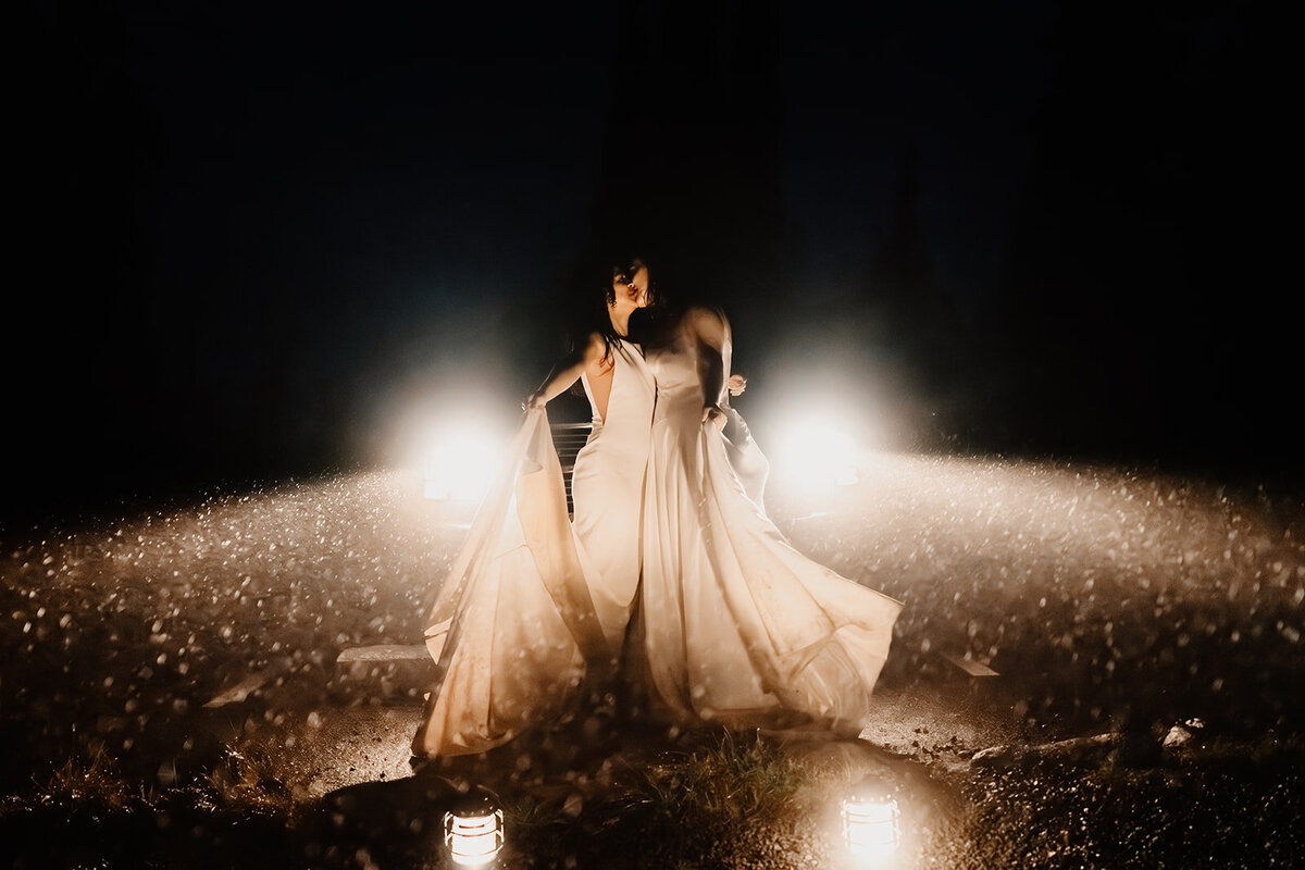 two brides in wedding dresses dance in front of the headlights of their adventure elopement photographers truck. the rain sparkles in the headlights. 