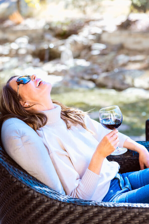 Girl in sunglasses throws her head back and laughs while holding a glass of wine.