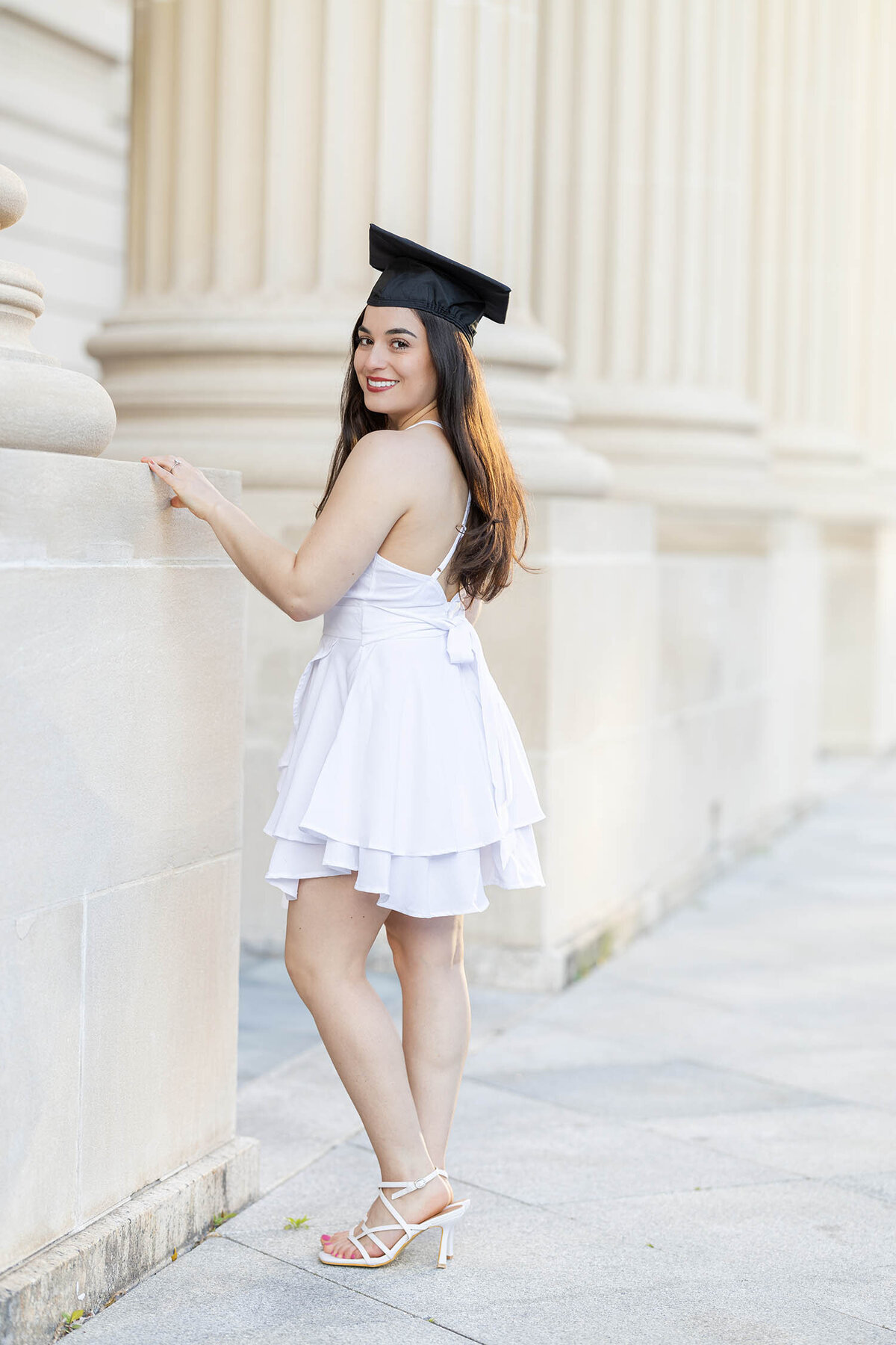 Graduate girl wearing a white dress poses to the side at golden hour among large cream color columns on Yale University iconic campus