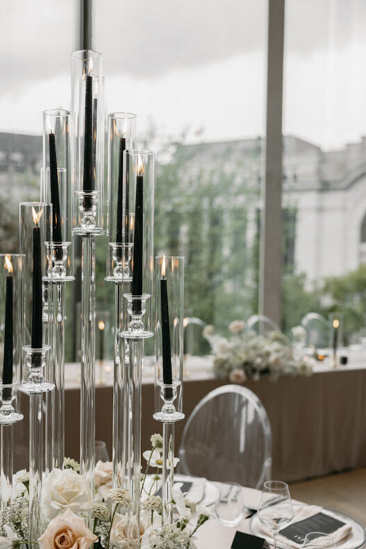 A modern glass candelabra with lit black tapered candles serves as a stylish centerpiece at a wedding in the National Arts Centre's O'Born Room, complemented by taupe linens and clear acrylic chairs.