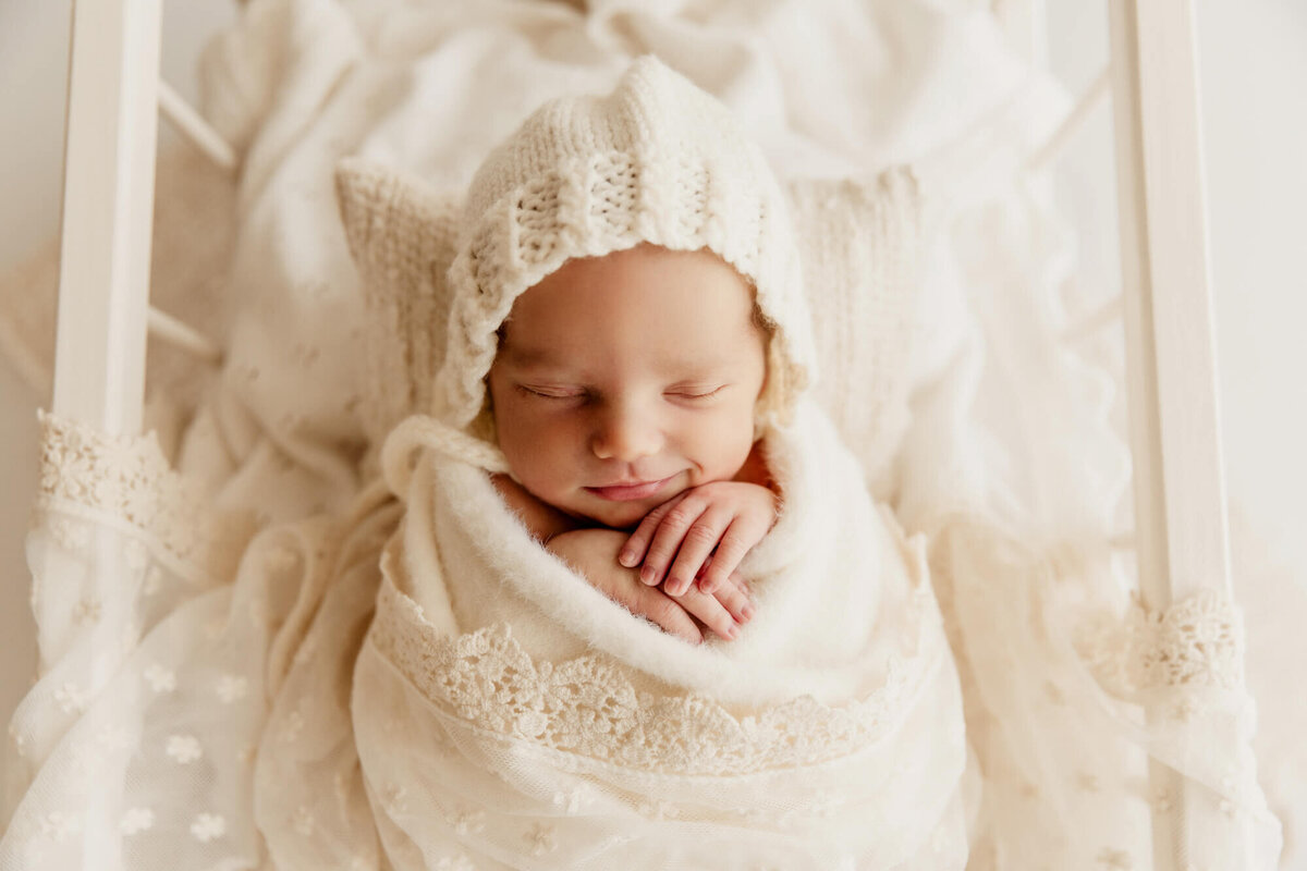 Smiling baby girl with her hands under her chin sleeping and wearing a white knit bonnet.