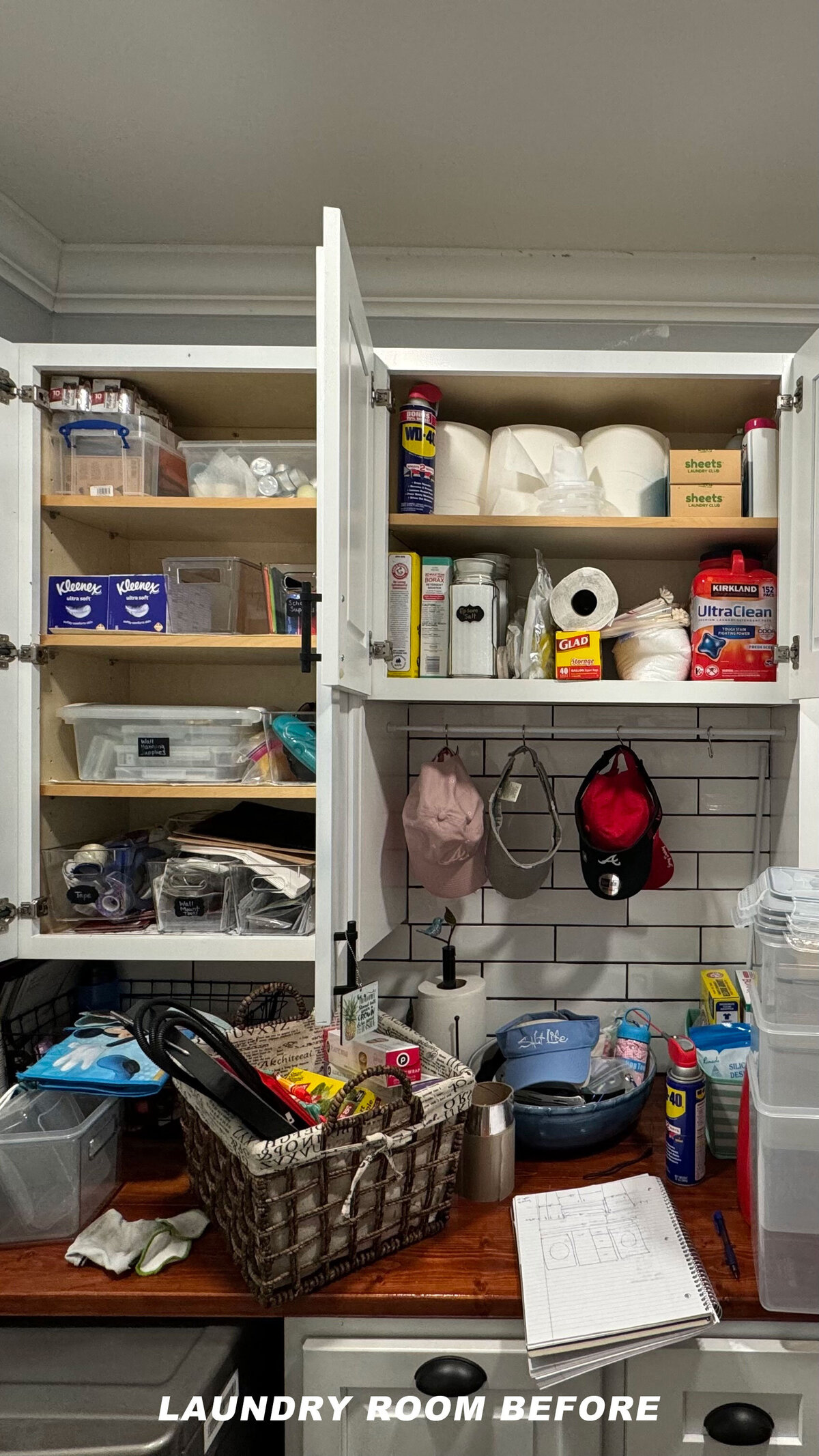 A cluttered laundry room with cabinets filled with various items like toilet paper, cleaning supplies, and storage boxes. Hats hang from hooks on the backsplash. A basket and a notebook are on the countertop. Text reads "LAUNDRY ROOM BEFORE.