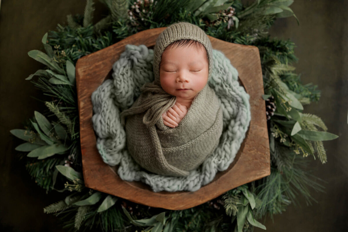 Newborn baby wrapped in a green knit blanket and hat, peacefully sleeping in a wooden bowl surrounded by lush greenery.