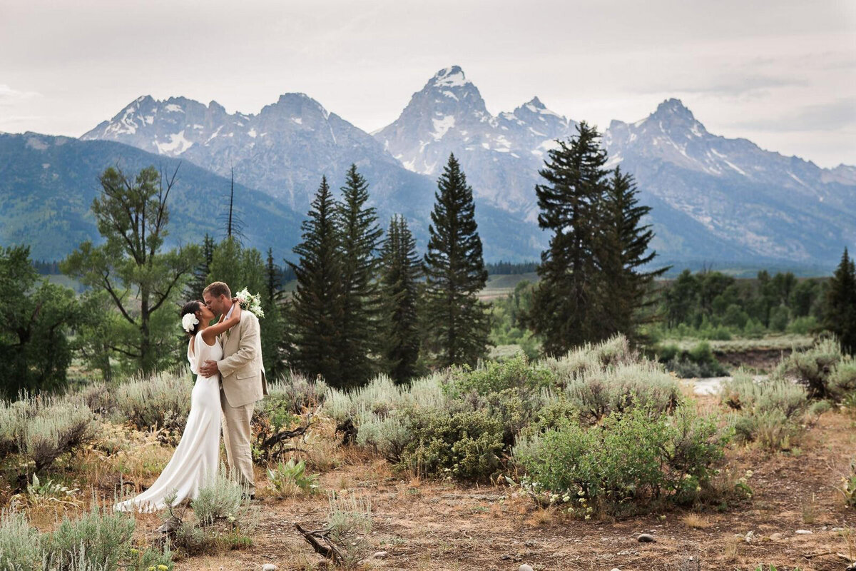 A bride and groom kiss among a field of sagebrush backdropped by the soaring peaks of the Teton range in Grand Teton National Park