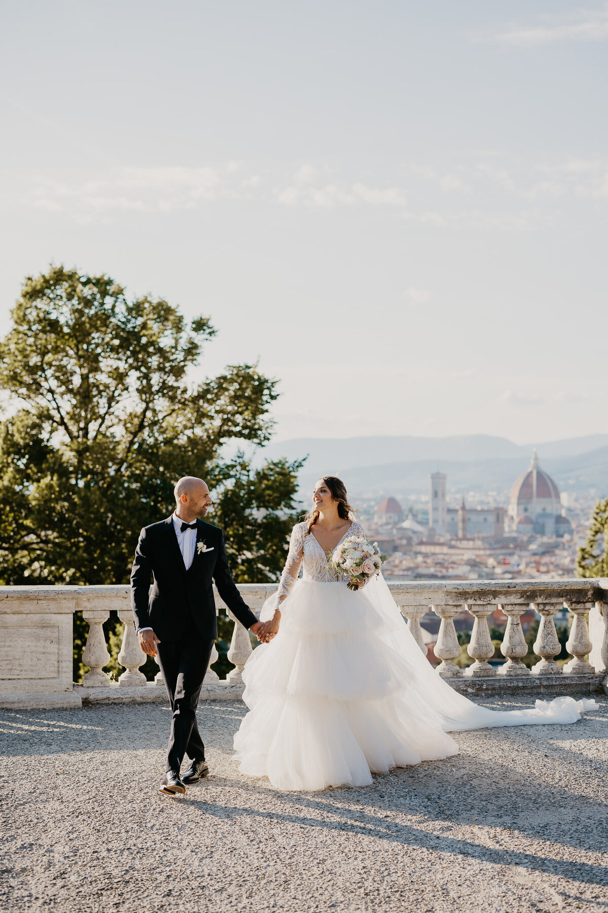 Bride and groom walking hand in hand with Florence skyline view from San Miniato al Monte, wedding photographer Tuscany.
