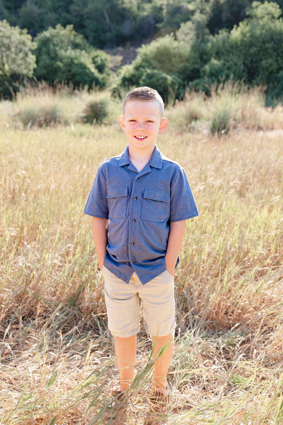 A young boy in a blue button-up shirt and khaki shorts stands confidently in a field of tall grass. His bright expression and relaxed posture capture a classic childhood portrait.