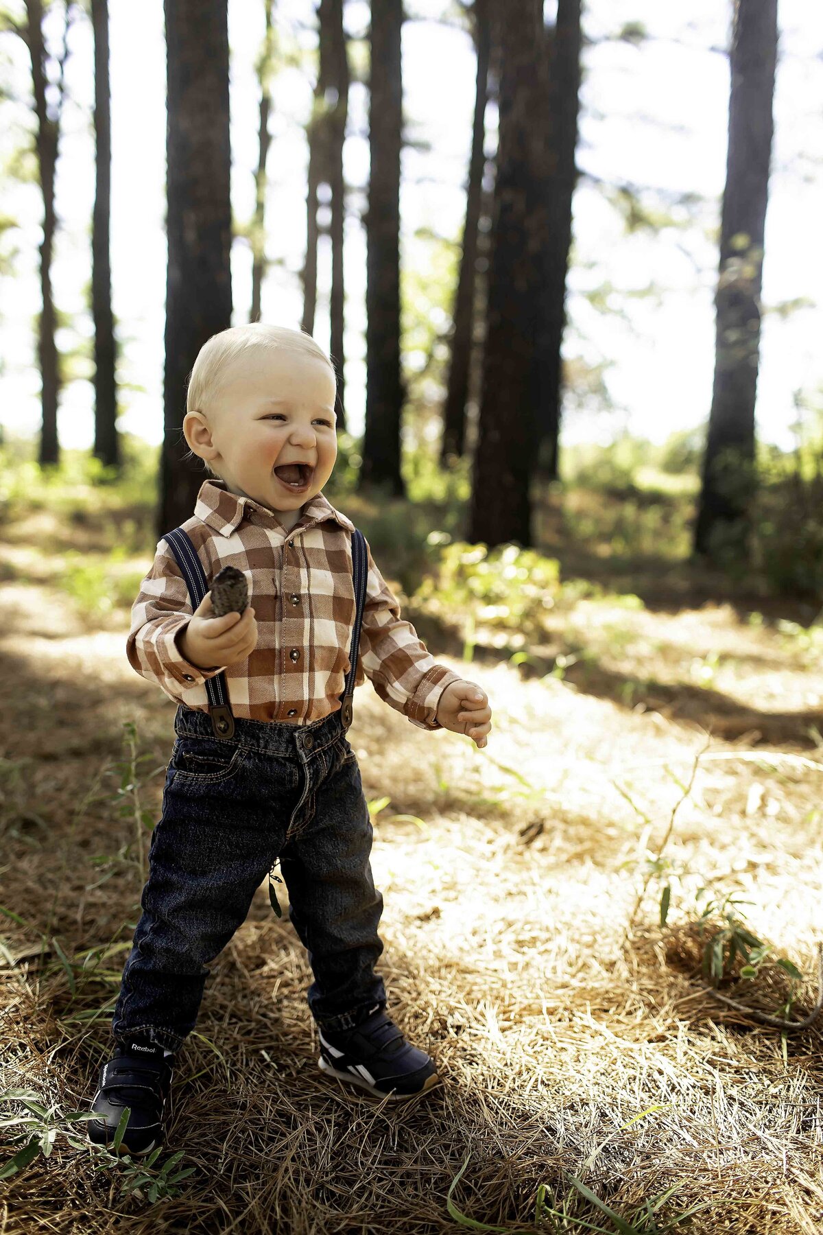Birthday boy wearing suspenders and overalls, looking cheerful while standing among the lush pine trees