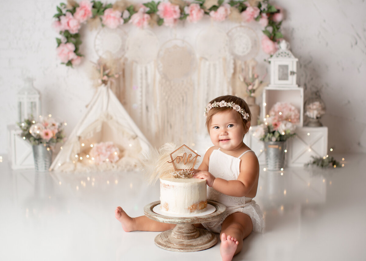 Pink and white themed cake smash. Baby girl in white outfit is sitting behind a white cake with gold "one" toppers. In the background there are boho macrame wall hangings and a flower garland.
