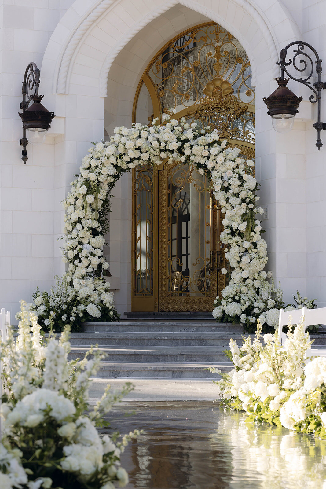 A grand wedding archway made of white roses, framing golden ornate gates of a castle.