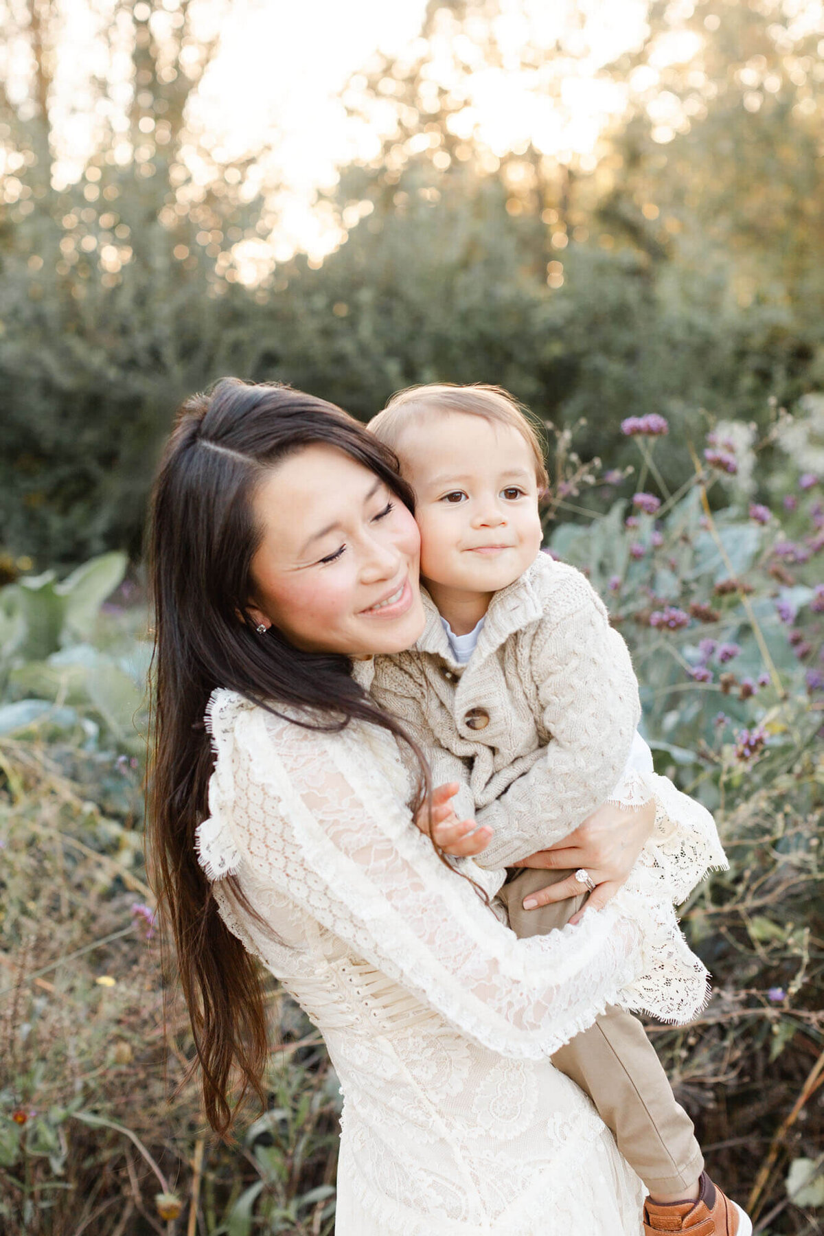 Gorgeous Mom with long dark hair cascading down her back and wearing a gorgeous Zimmerman dress holding her toddler son in khaki and cream. They are cheek to cheek with a treeline behind them as the sun is setting and creating a beautiful golden glow.