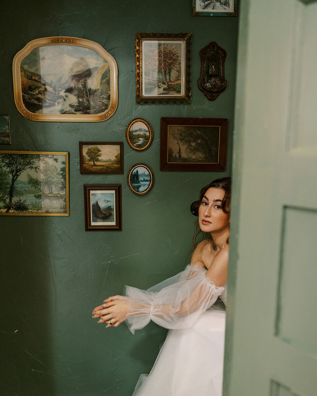 A woman sits in a wedding dress in front of a green wall with photos. 
