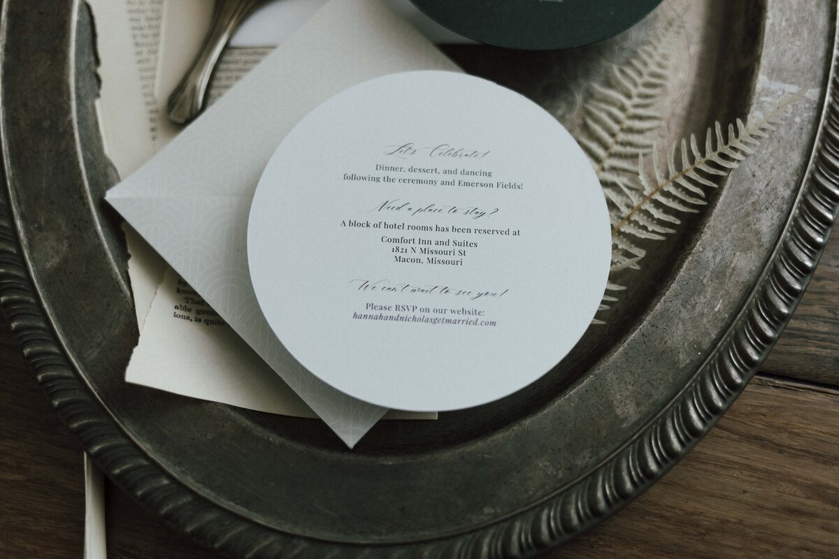 White rounded wedding invitation with black cursive font and an ivory and white textured square envelope atop silver tray.