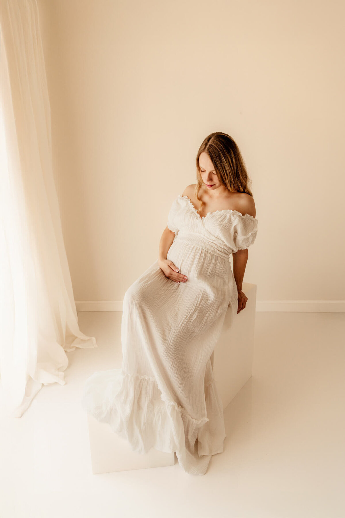 Woman with white off the shoulder sweetheart dress sitting on a white block while looking down at her baby bump for an OKC maternity photo session.