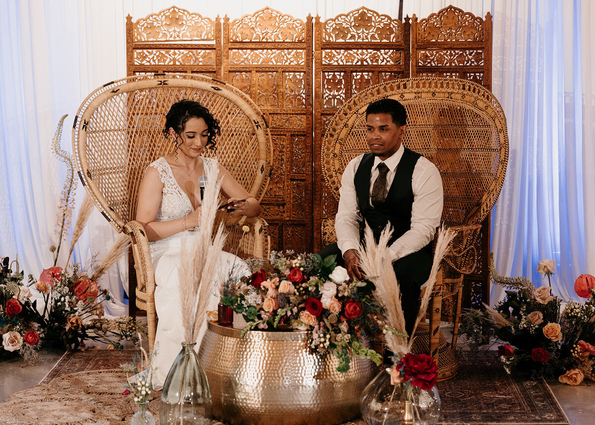 Bride and groom seated on wicker peacock chairs, surrounded by flowers and greenery, creating a boho-chic atmosphere for their reception.