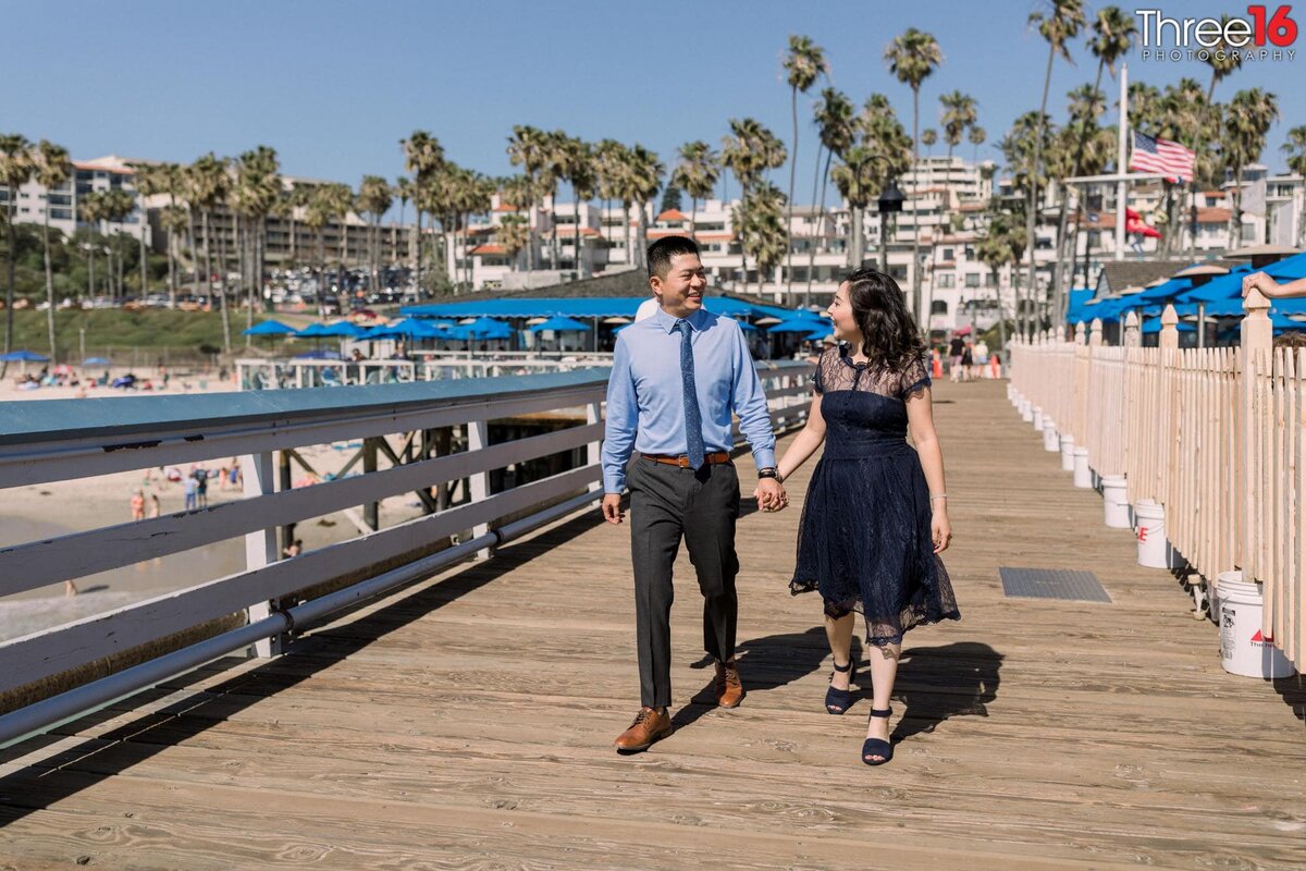 Engaged couple walk along the San Clemente Pier holding hands and talking with the city in the background