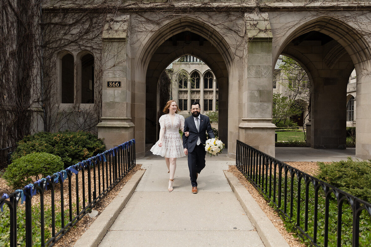 Happy couple walks quickly outside of church, They're smiling and their arms are linked, groom carries the bouquet. Both of them look at camera.