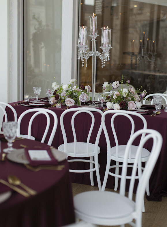 An elegant guest table at a National Arts Centre wedding, featuring a tall glass candelabra with blush tapered candles, burgundy satin linens, and low blush and burgundy floral centrepieces on each side of the candelabra. The table surrounded by white Bentwood guest chairs.