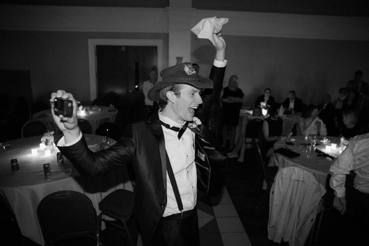 Groom candidly waves napkin above head while snapping a photo with his other hand at his wedding reception.