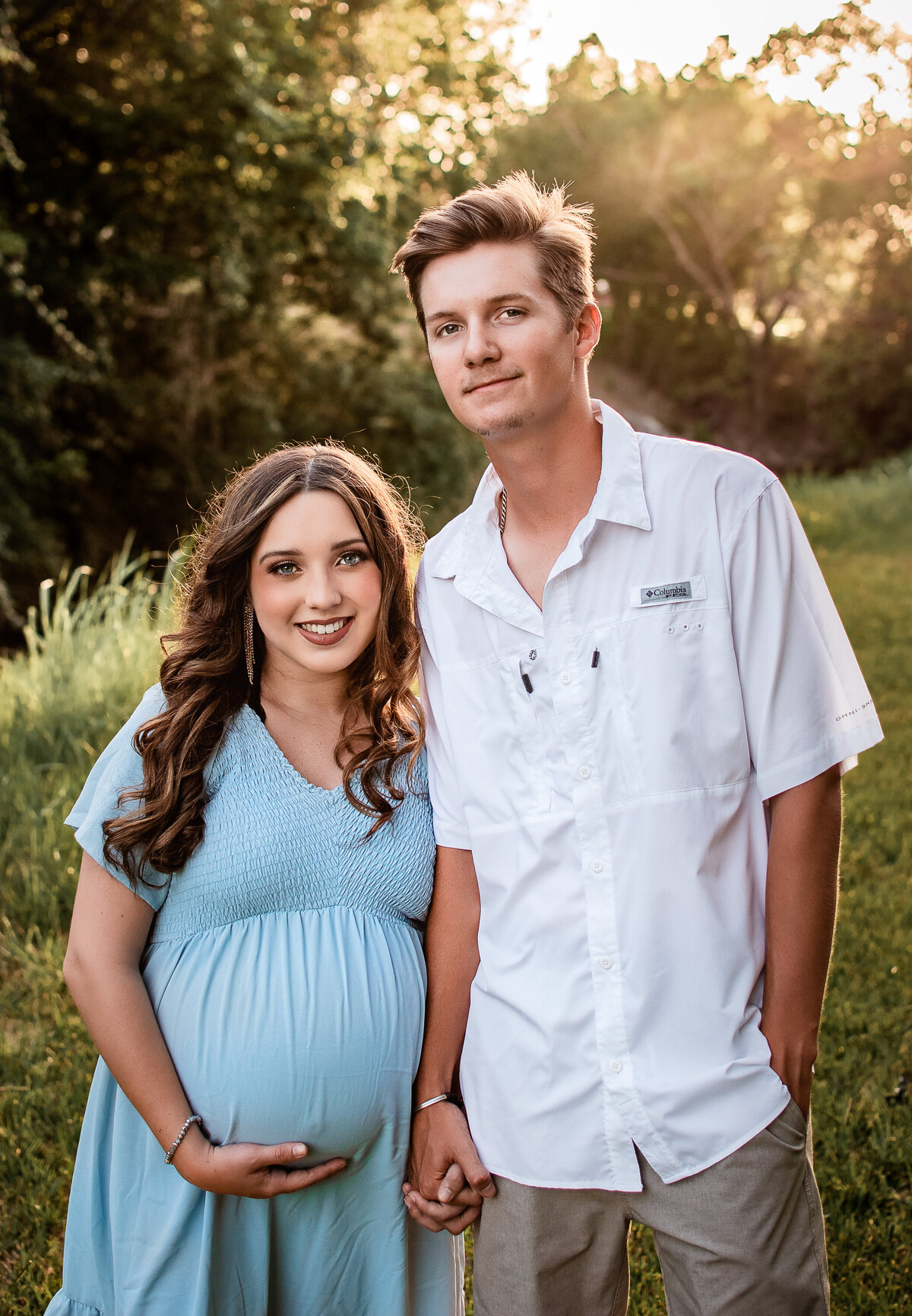 A pregnant couple holds hands and smiles at the camera.
